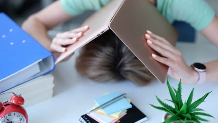 Woman with head on table and laptop covering her head.